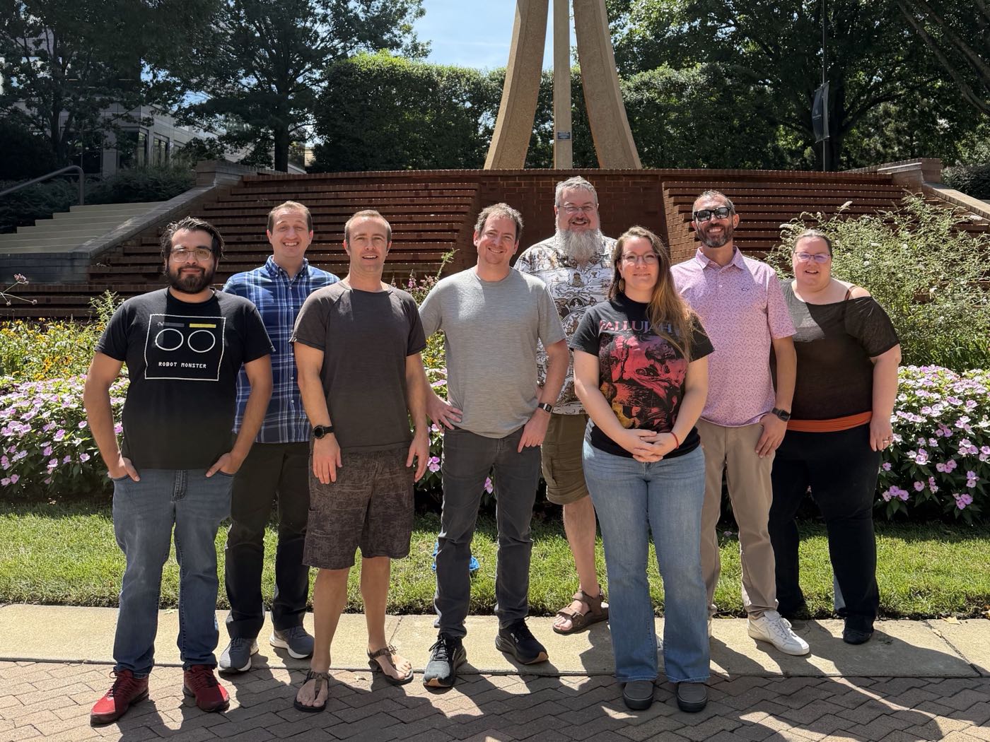 The TruBase team, eight people, standing together on a sunny day in front of a brick amphitheater and carillon tower.